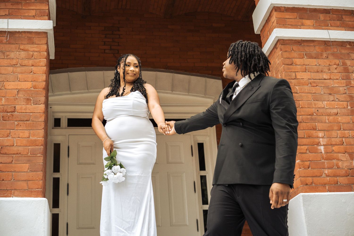 Bride and groom hold hands during outdoor ceremony in front of brick building with white doors.