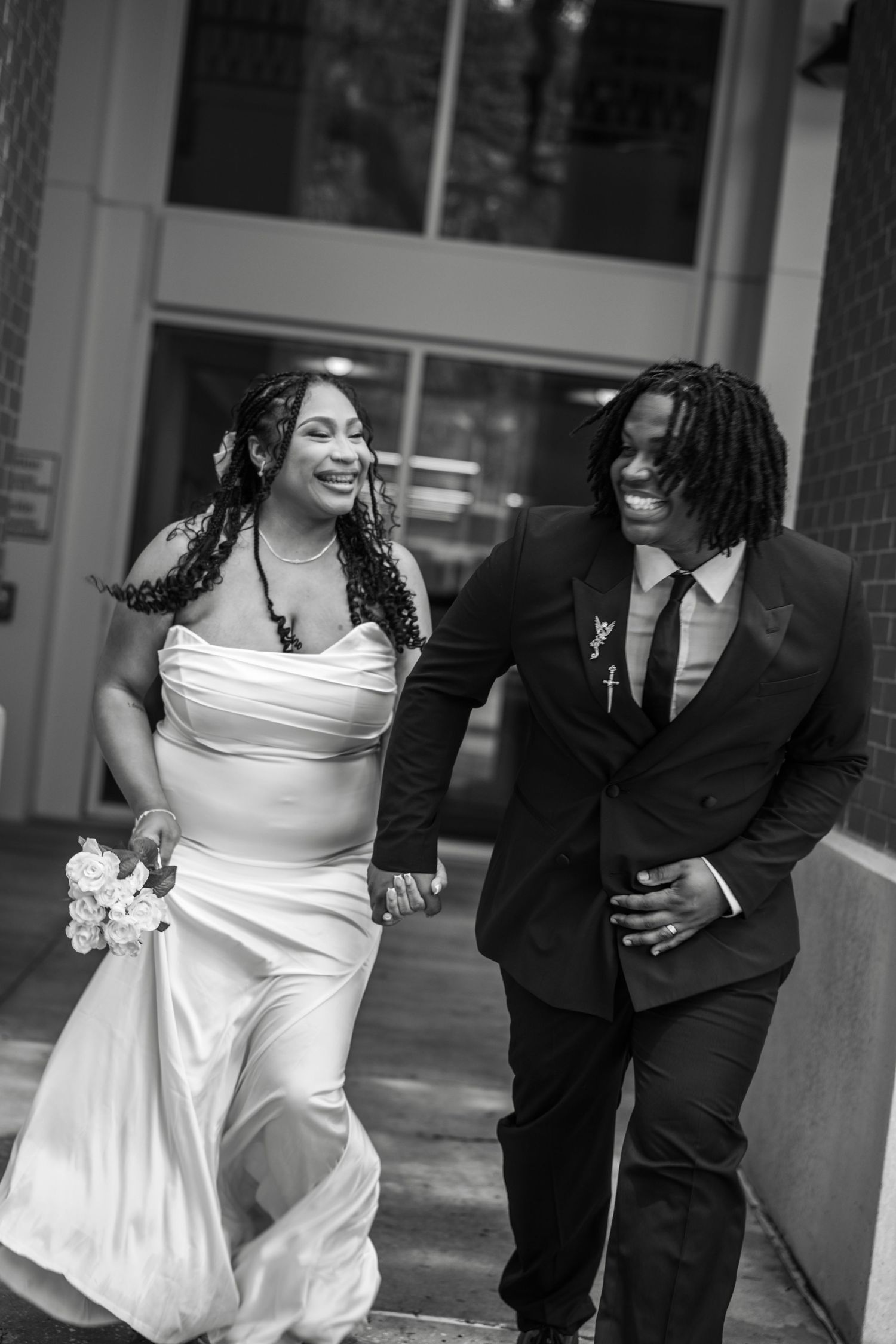A playful black and white candid moment of a bride in a white dress and groom in black suit walking and laughing together.
