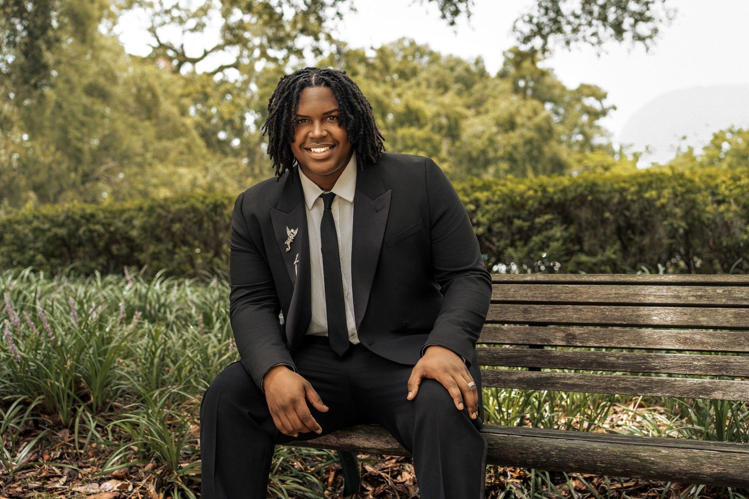 A person in a black suit sits smiling on a wooden bench in a scenic outdoor garden setting.