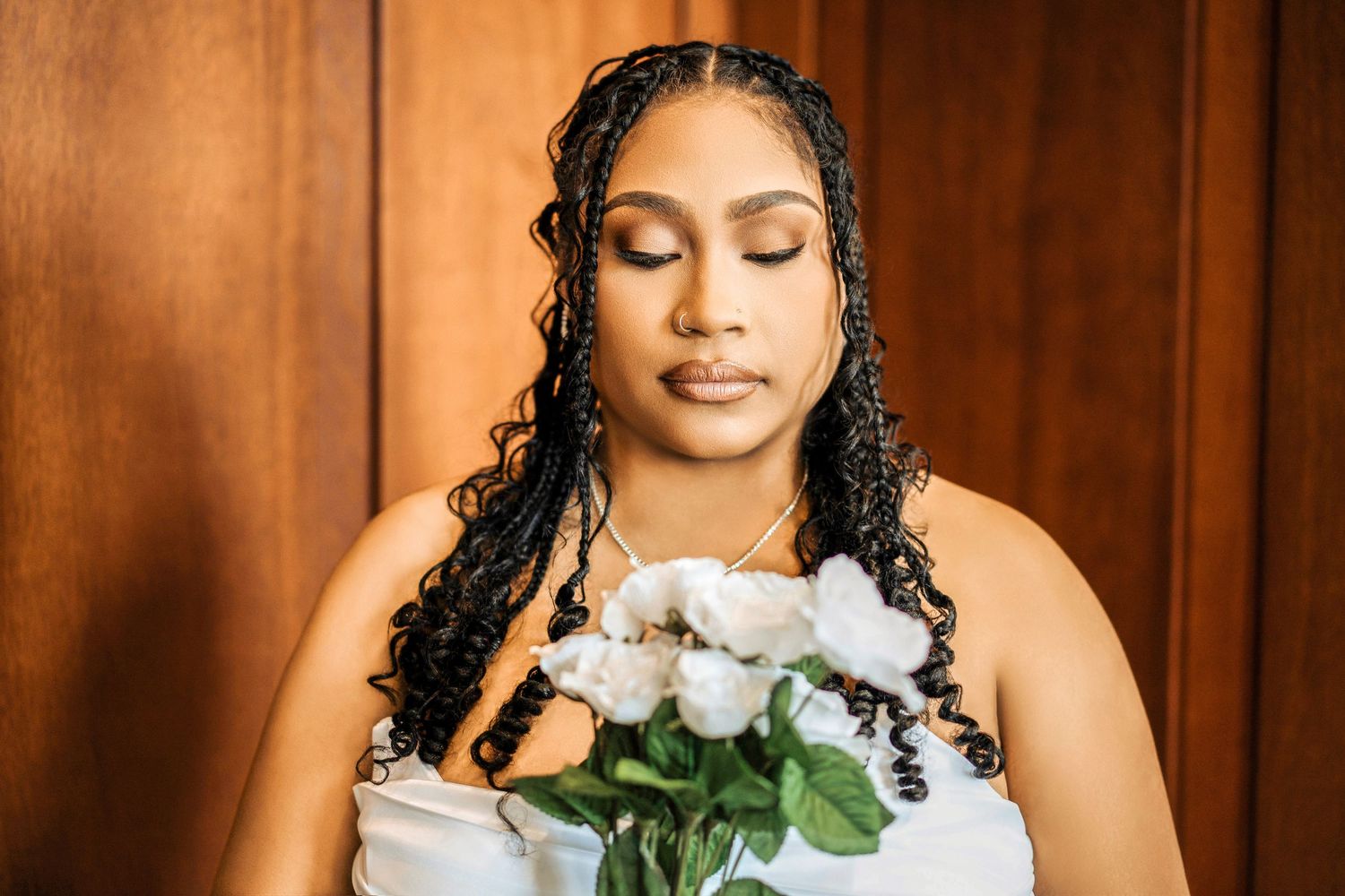 Beautiful bride in white wedding dress holds bouquet of white roses against brown wooden background.
