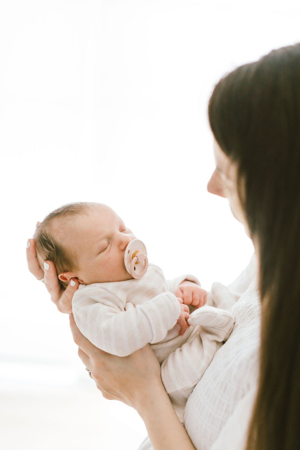 Parent holds sleeping baby with pacifier in front of bright window.