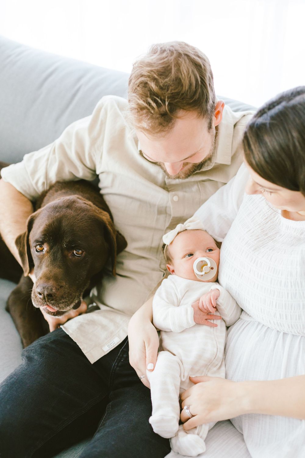 Chocolate labrador joins intimate family portrait session with newborn on white couch.