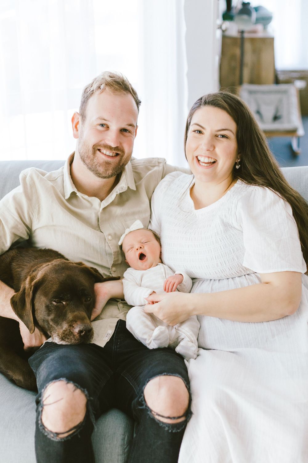 Family relaxes on couch with their newborn baby and chocolate lab dog in a bright living room.