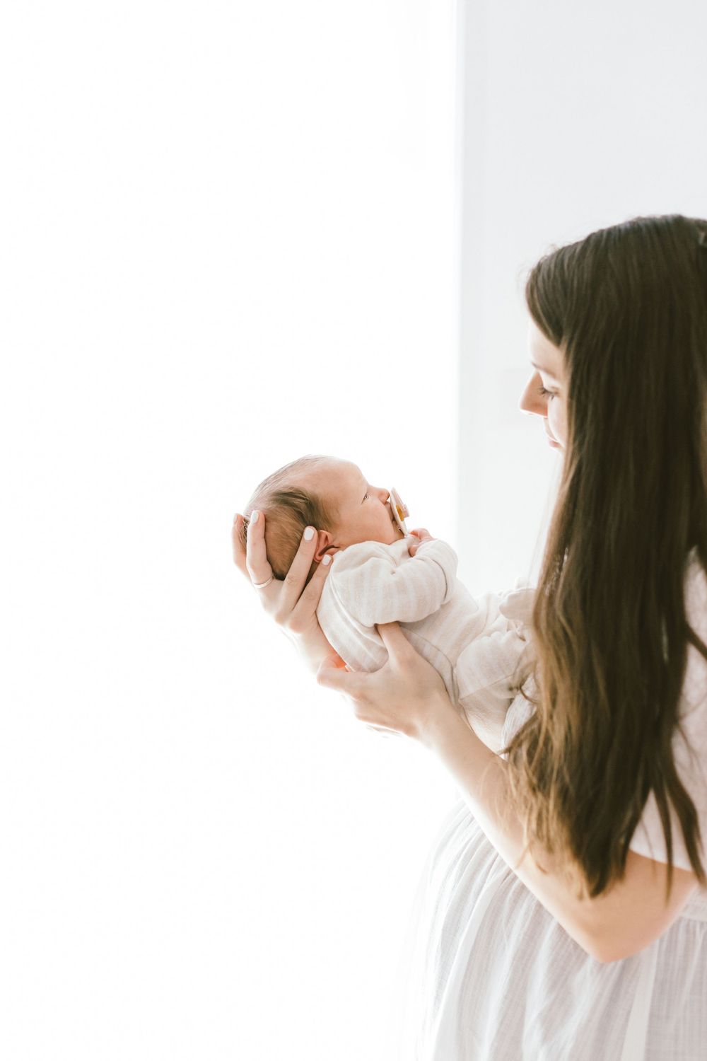 Woman in white dress holds sleeping newborn baby against bright window backdrop.