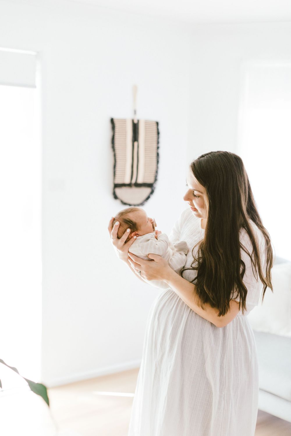 Woman in flowing white dress cradles newborn baby in bright minimalist room.