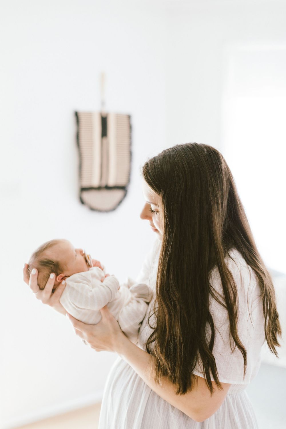 Woman in white dress holds infant while standing in bright room with wall decor.