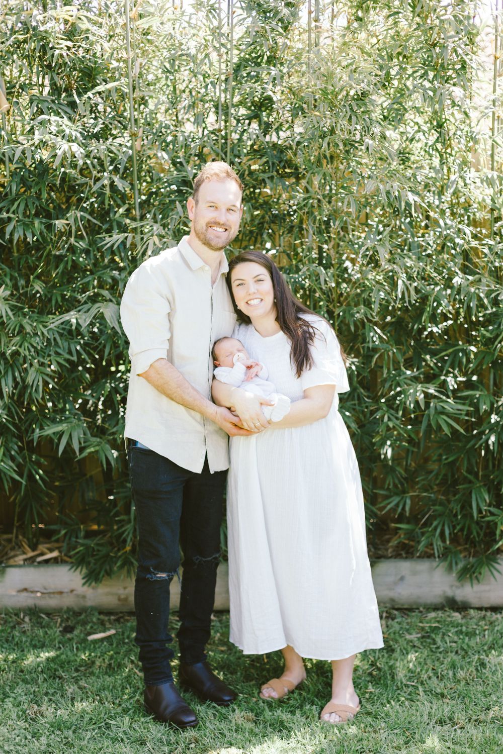A couple holds their newborn baby while standing in front of lush bamboo plants on a sunny day.