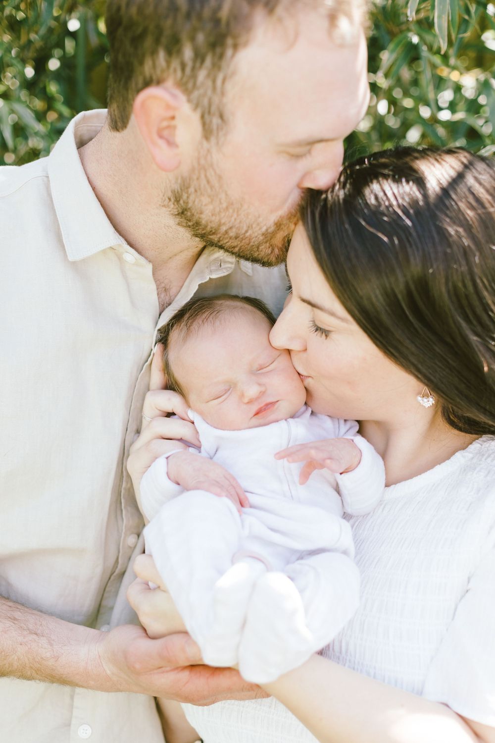 Parents sharing an intimate moment with their sleeping newborn baby dressed in white.