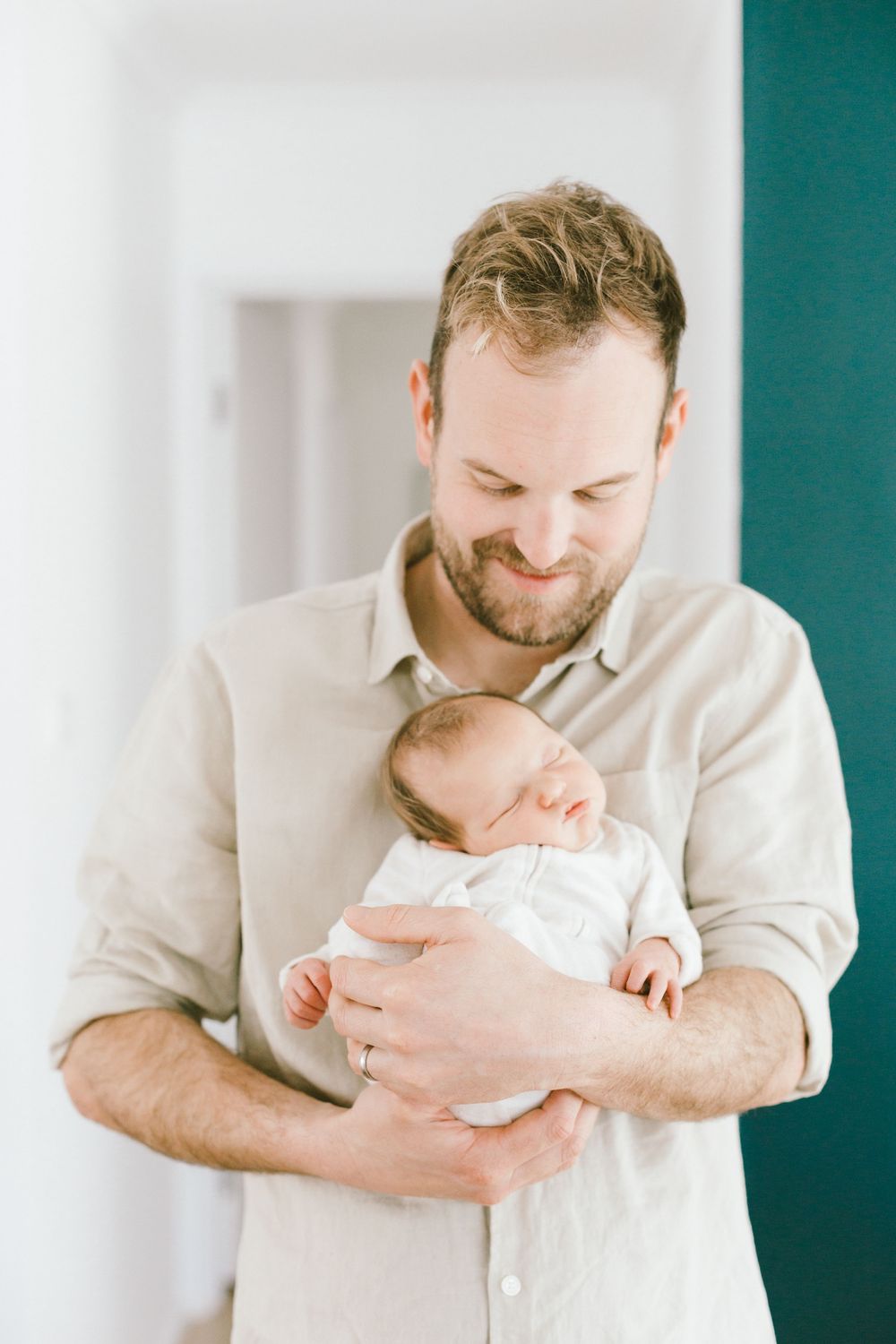 Parent in beige shirt holding sleeping newborn against teal and white wall.
