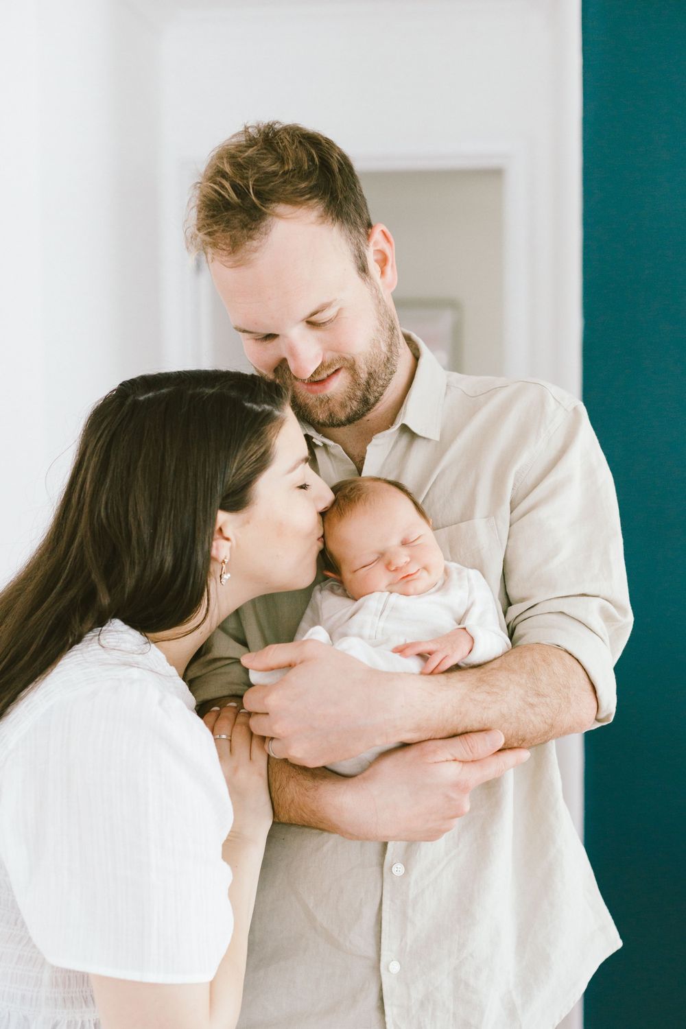 Parents embrace while holding their sleeping newborn baby against teal wall.