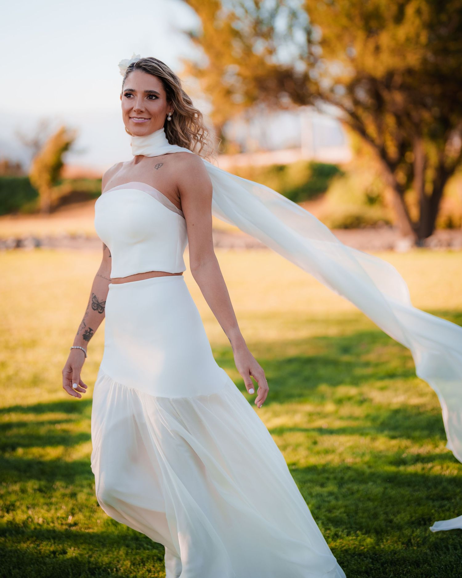 Radiant bride in white two-piece wedding dress with flowing veil twirls on green grass at sunset.