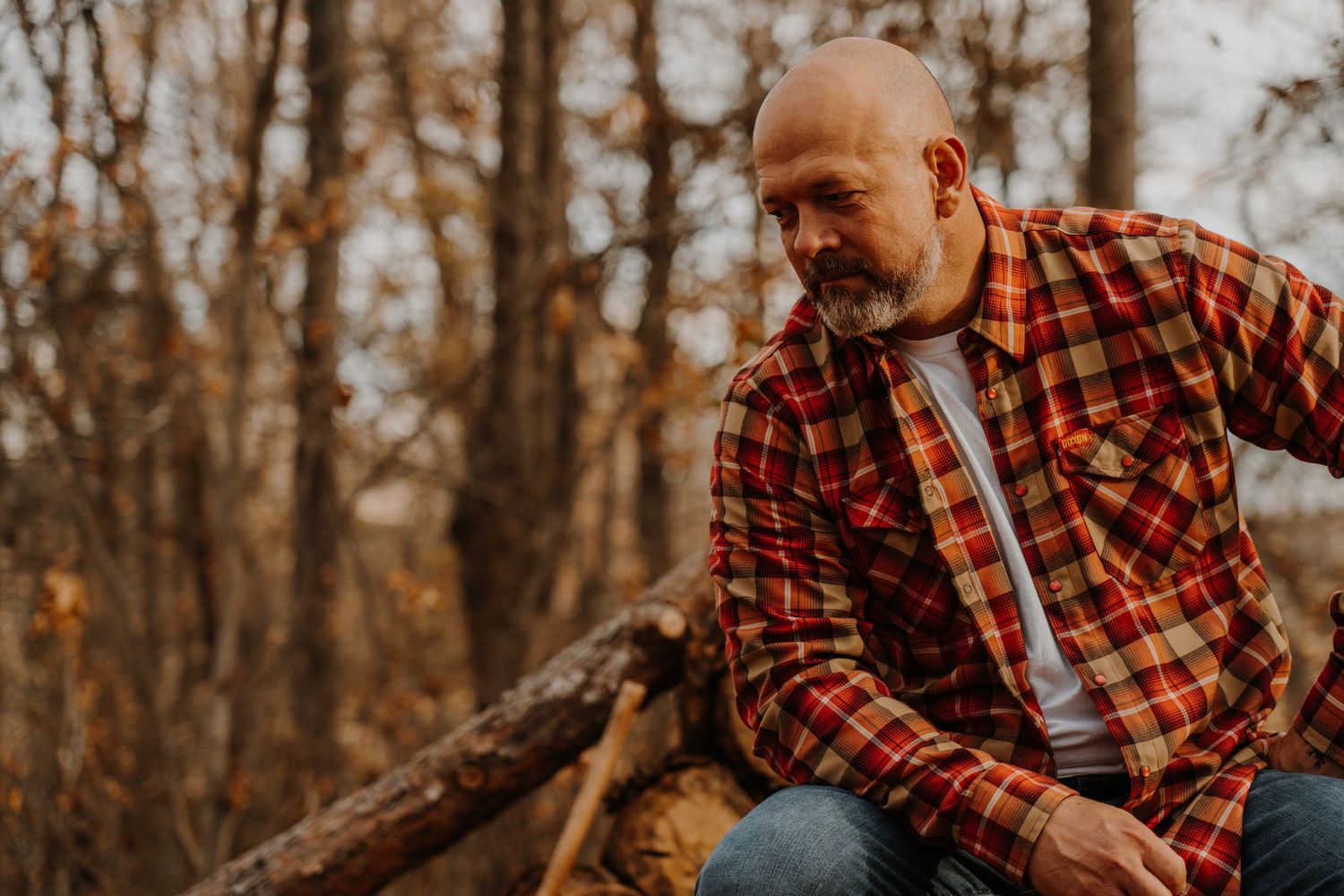 Person in red plaid shirt sitting outdoors during autumn season with blurred forest background.