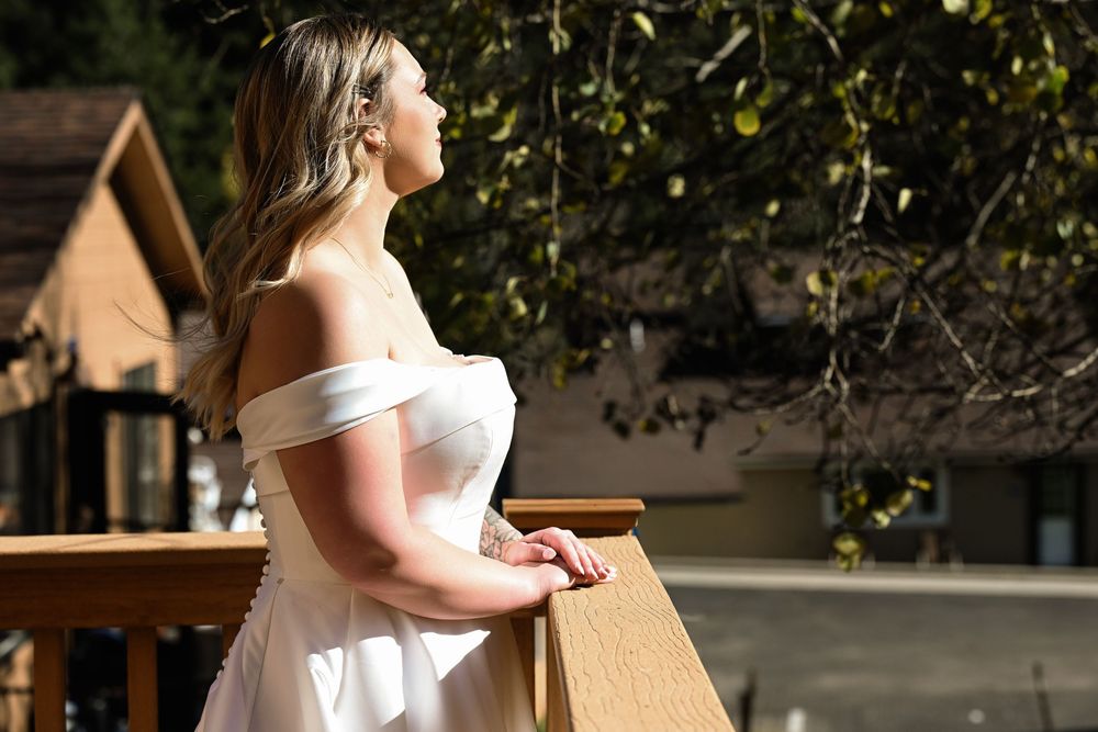 A person in a white off-shoulder dress stands on a balcony in warm sunlight with a wooden birdhouse visible nearby.