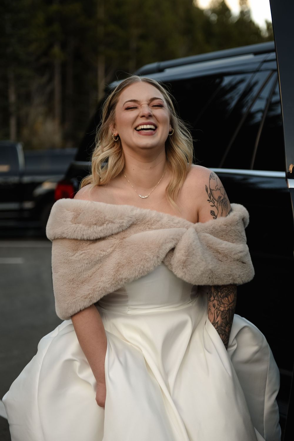 A bride in a white wedding gown with a plush off-shoulder fur wrap poses outdoors at dusk.