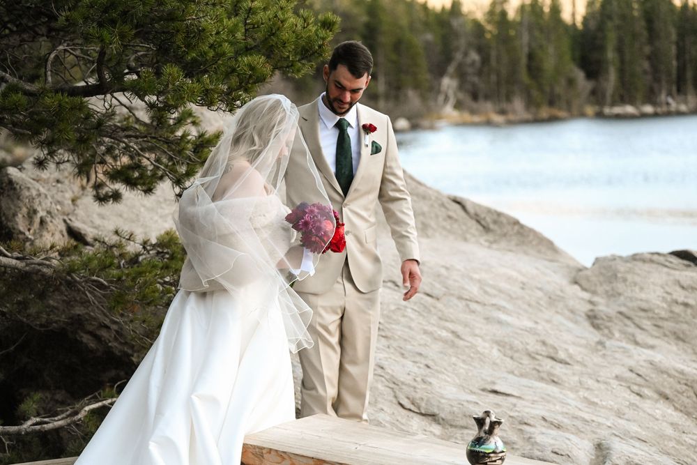A couple in wedding attire shares a romantic moment on a scenic beach with mountains and forest in the background.