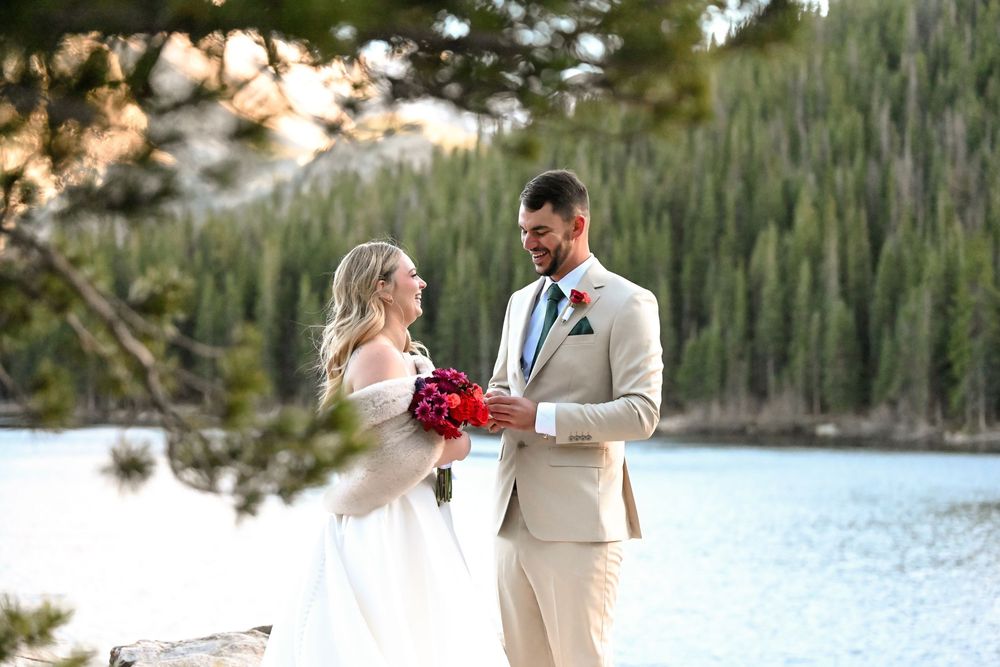 A couple in wedding attire stands by a serene mountain lake with evergreen forest in the background.