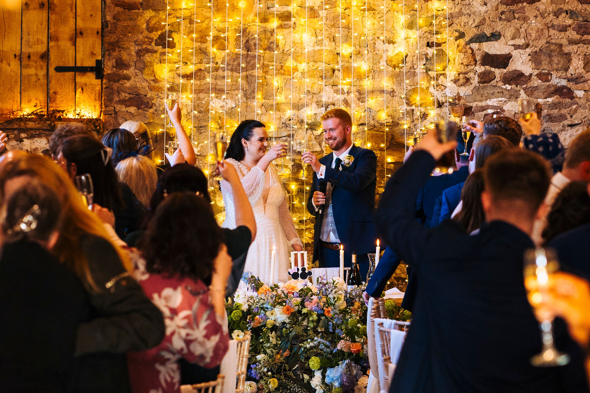 Wedding breakfast toast at Eden Barn in the lake district, couple cheers with fairy light wall behind them framed by the guests