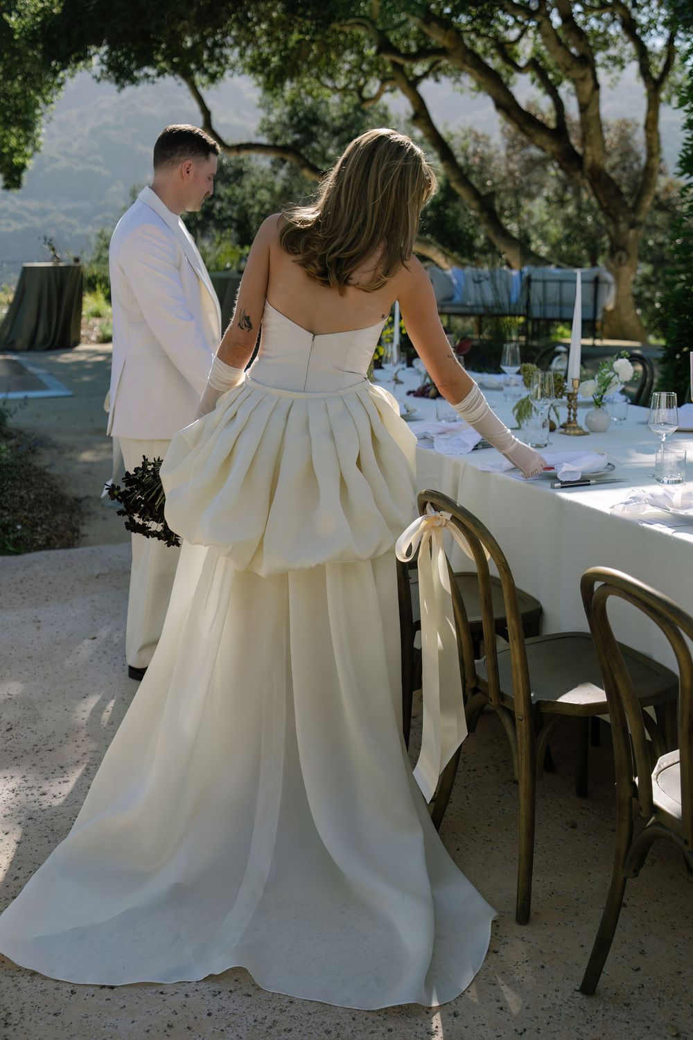 Couple in white attire walking through an outdoor wedding reception area with decorated tables.