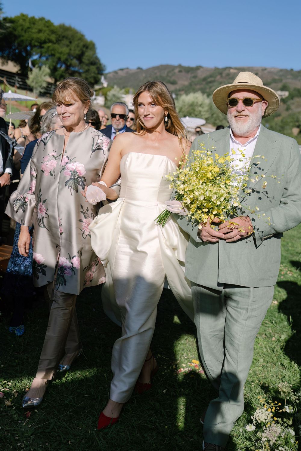 Wedding guests in formal attire gather outdoors on a grassy hillside holding yellow flower bouquets.