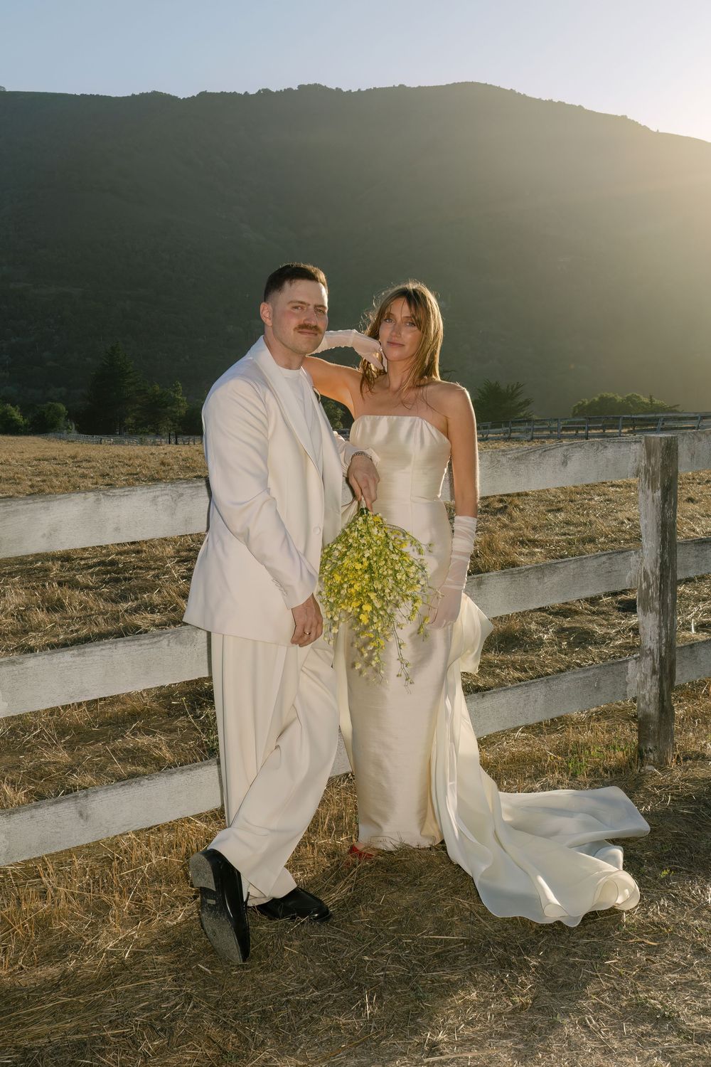 A wedding couple poses against a rustic wooden fence at sunset in a mountainous landscape captured by Danielle Conner Photography in Carmel