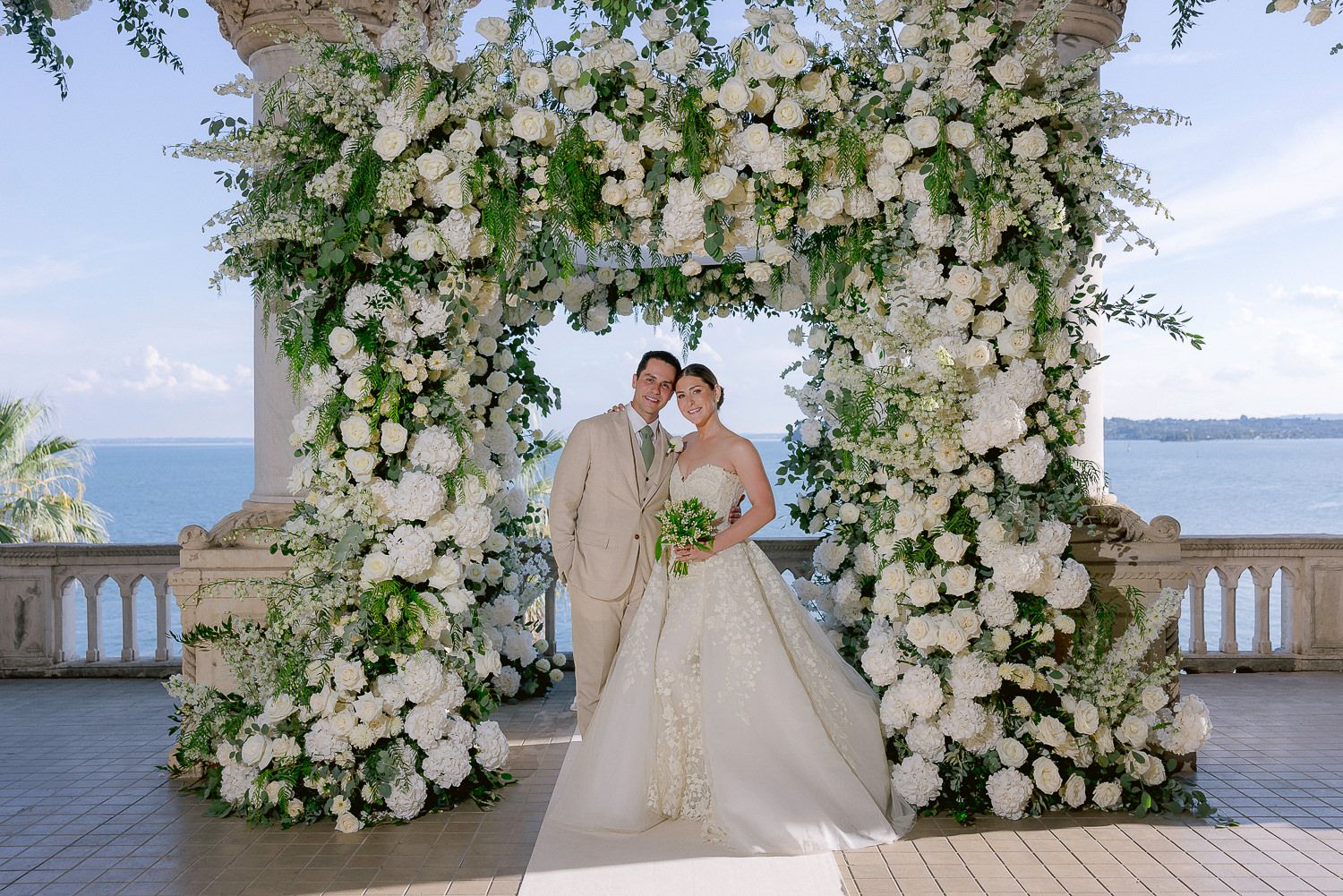 A stunning wedding photoshoot at a waterfront venue with elegant white floral arches and romantic coastal views.