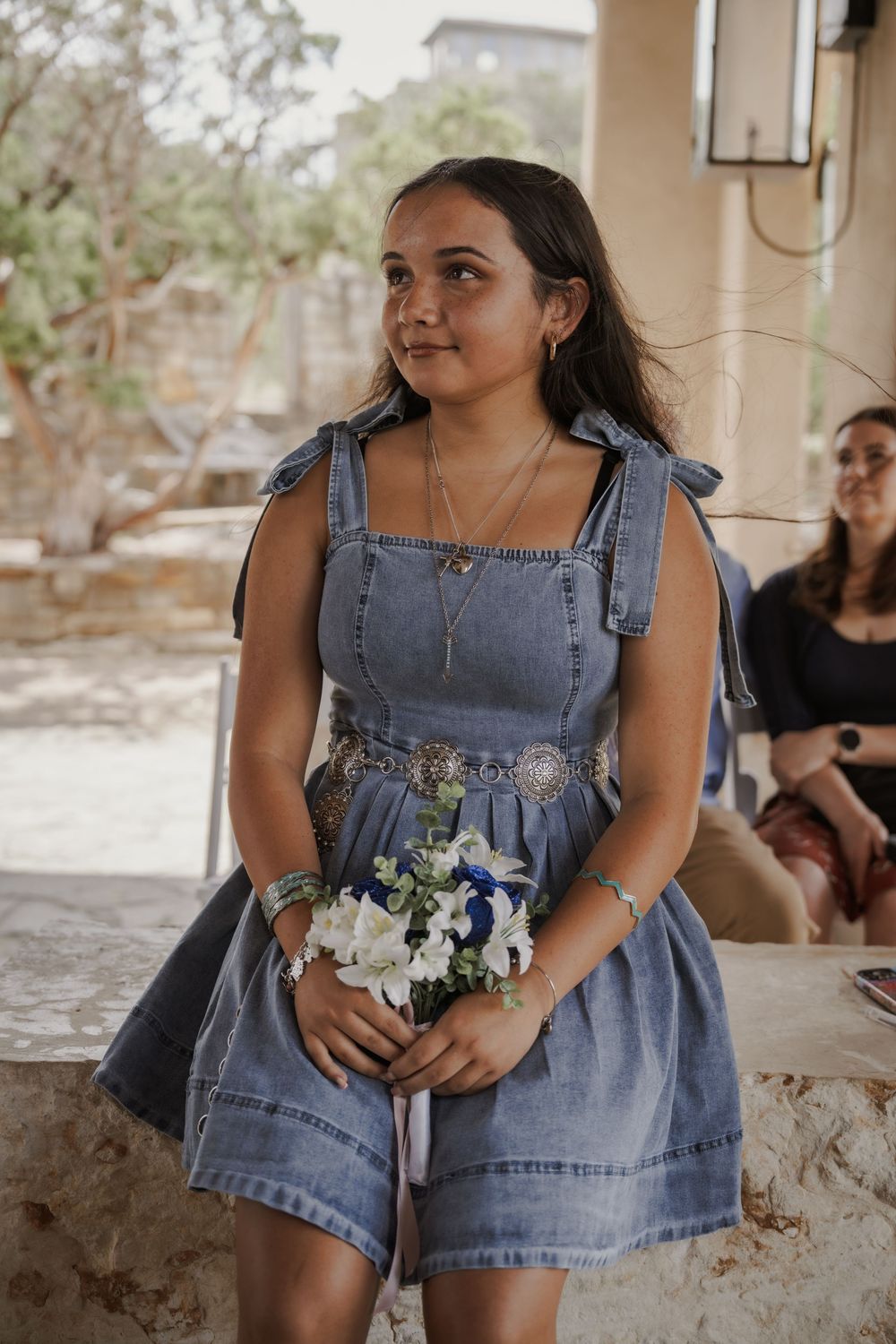Bridesmaid in denim dress holding small floral bouquet at outdoor wedding.