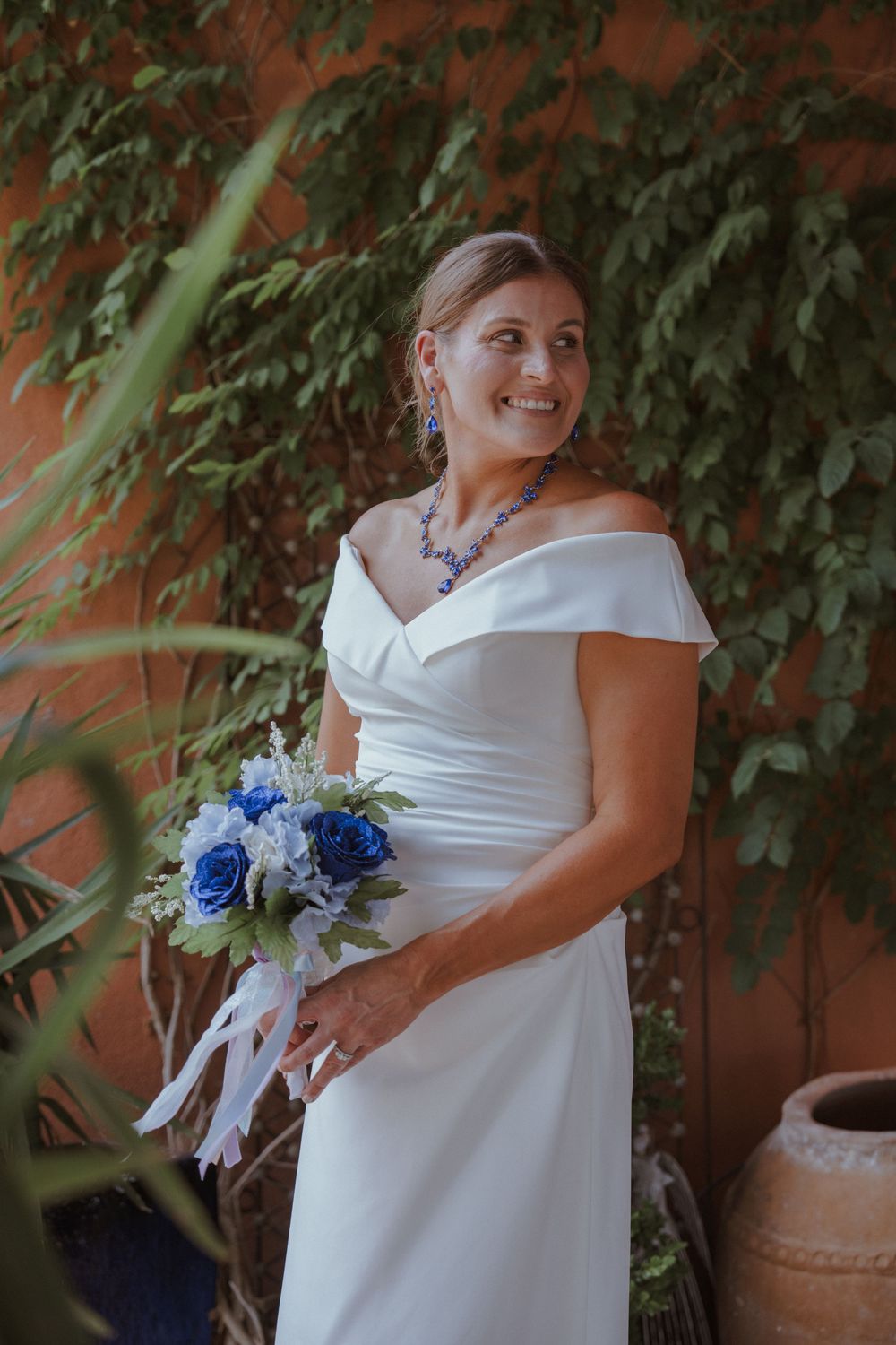 Bride in white dress holding blue and white floral bouquet against ivy-covered wall.