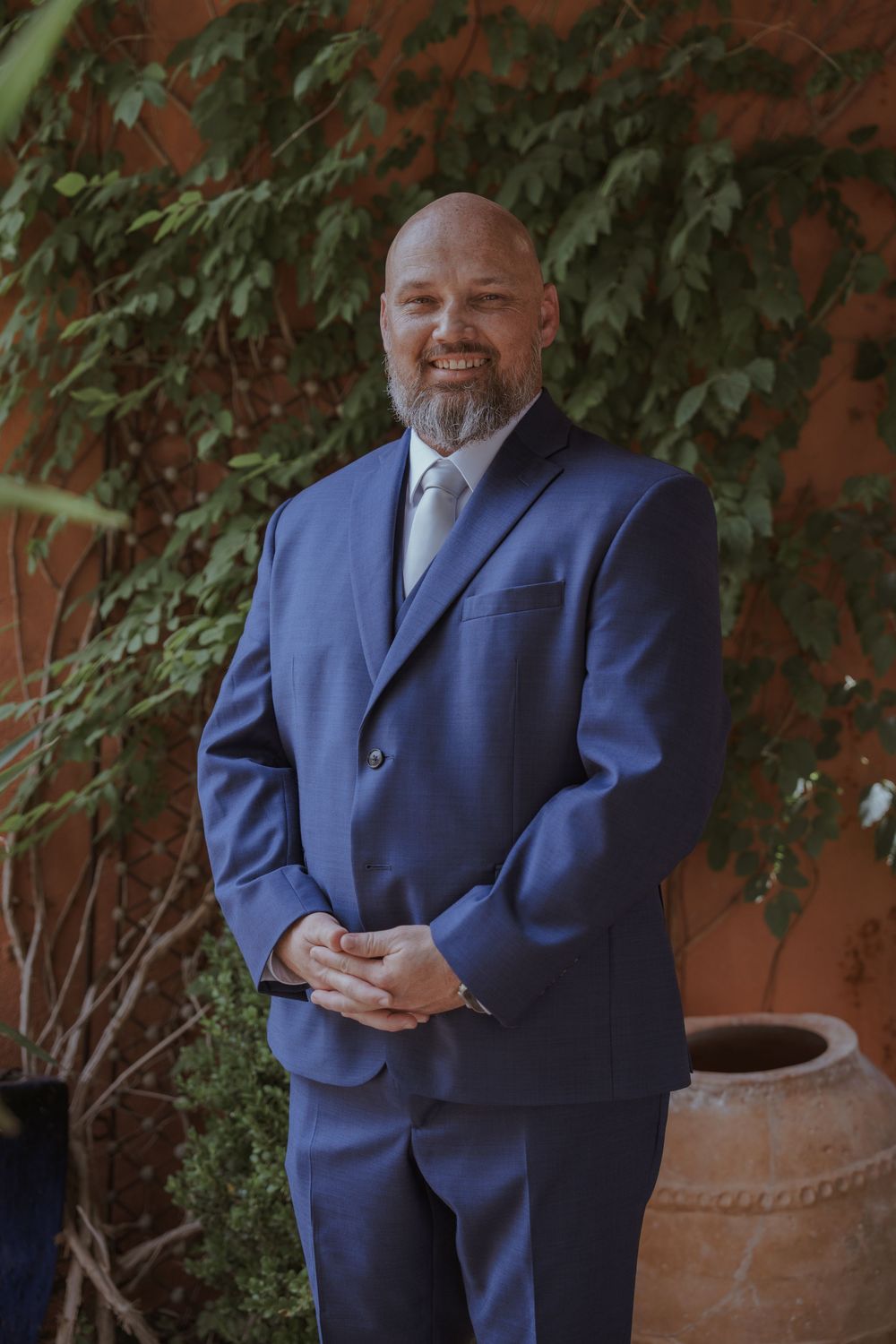 Groom in navy blue suit poses against terracotta wall with greenery.