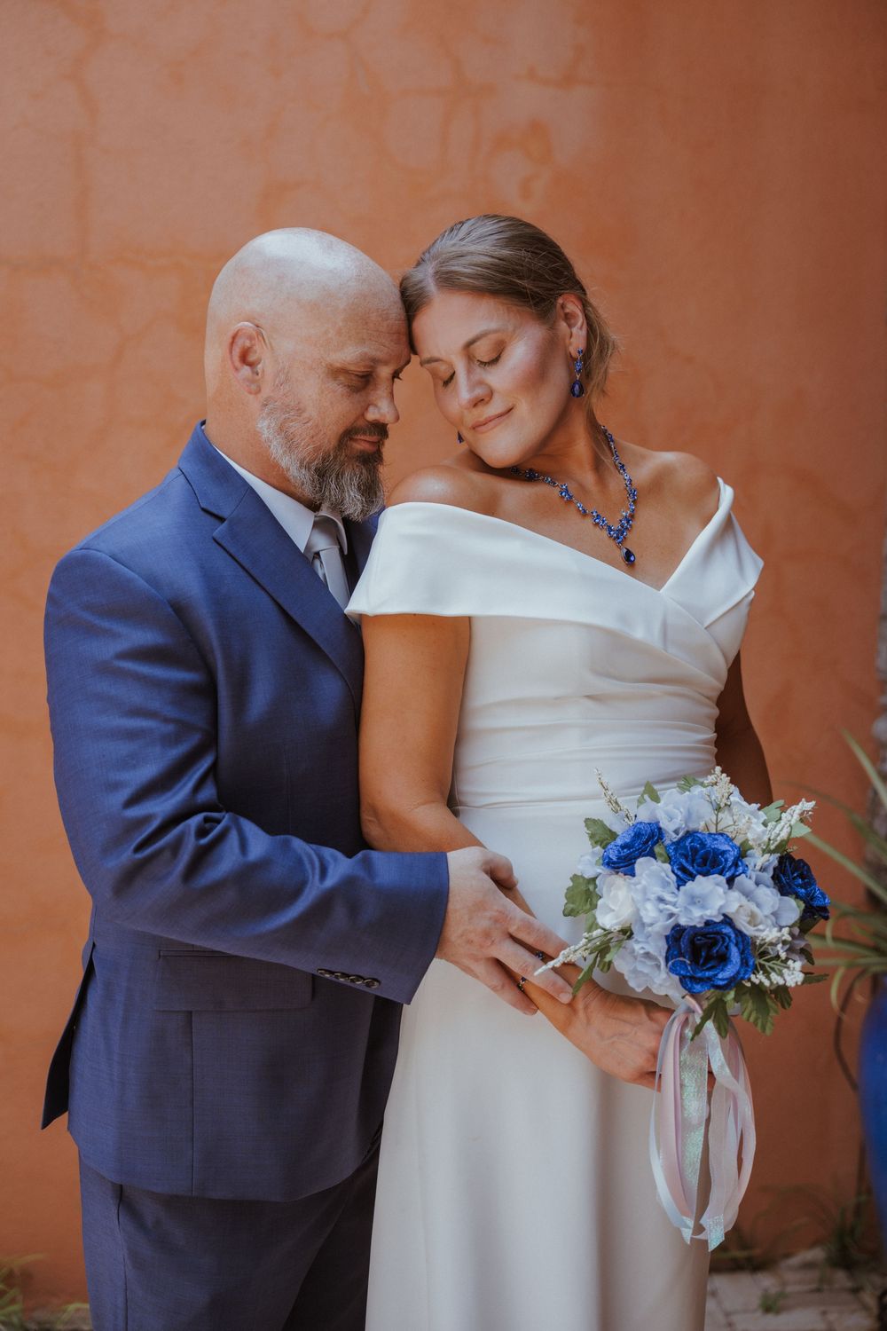 Intimate wedding moment captured against terracotta wall with blue floral bouquet details.