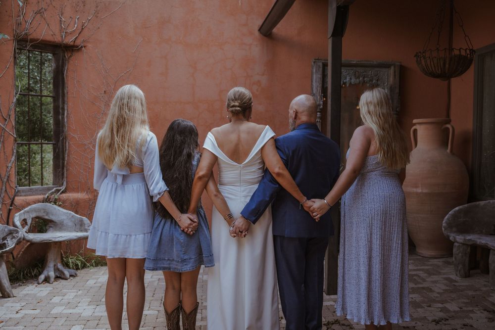 A group wearing matching lavender dresses stands together holding hands in a line against a rustic wall.