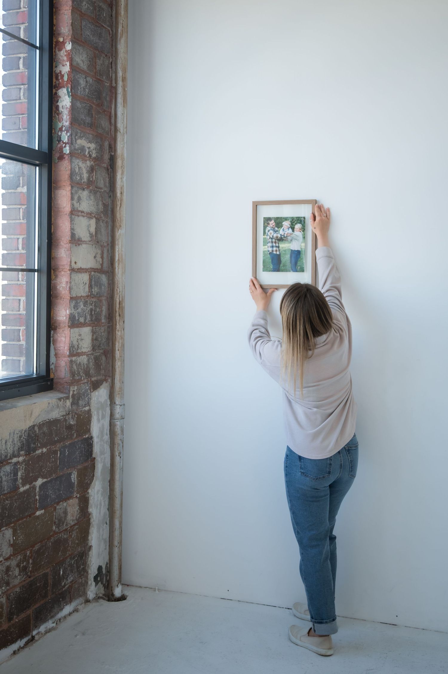 Person in jeans and gray shirt hangs pictures on white wall next to exposed brick in home renovation sequence.