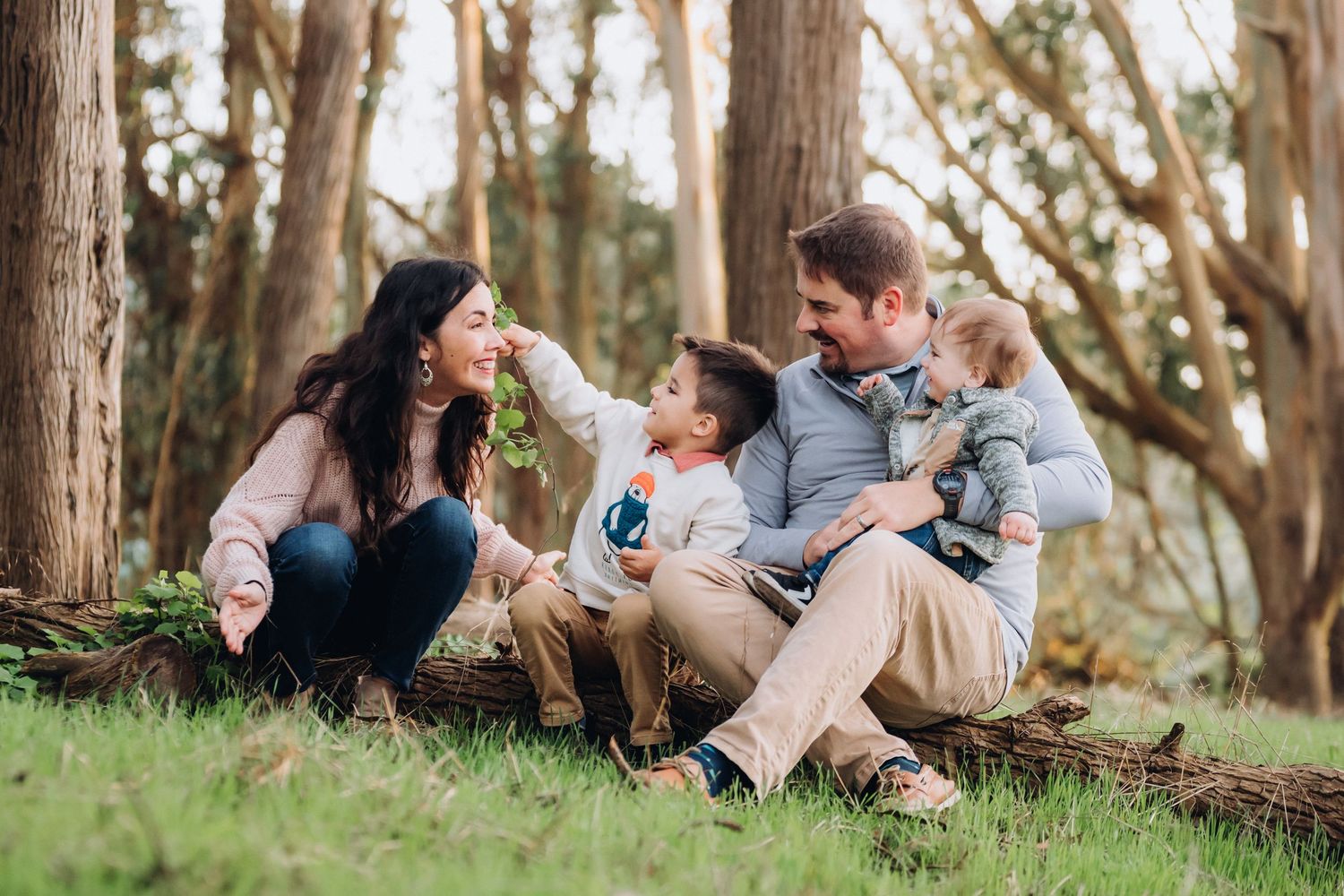 Family relaxes together on grass near trees during outdoor photo session in natural setting.