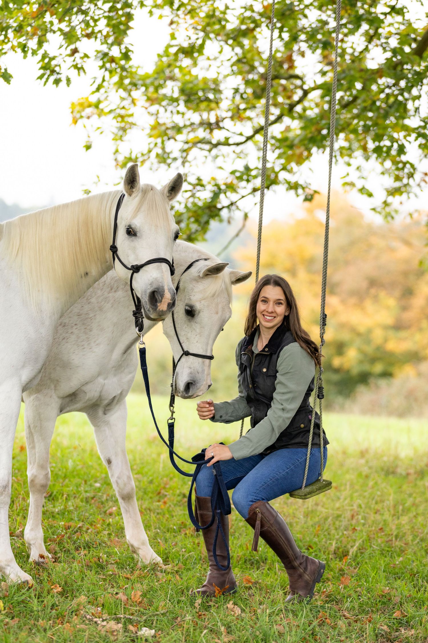 A brown horse stands next to a brick wall while someone interacts with it on an autumn day with fallen leaves.
