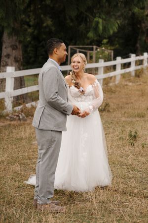 A couple in wedding attire dances together on a grassy hillside with a white fence in the background.