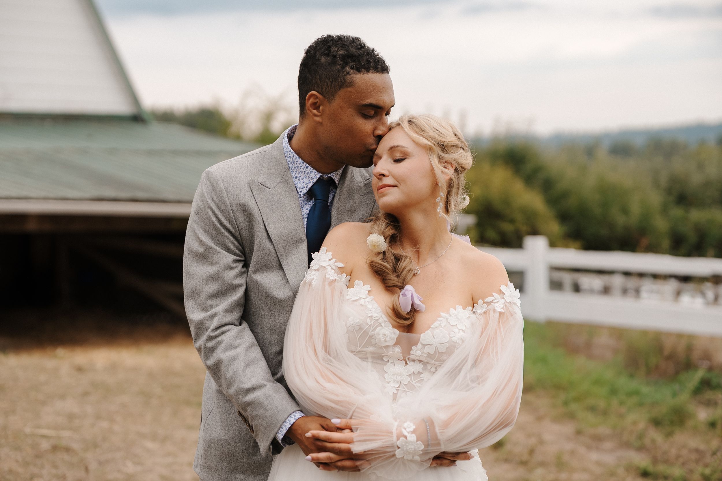 A romantic wedding couple embraces in a scenic outdoor setting with rolling hills in the background.