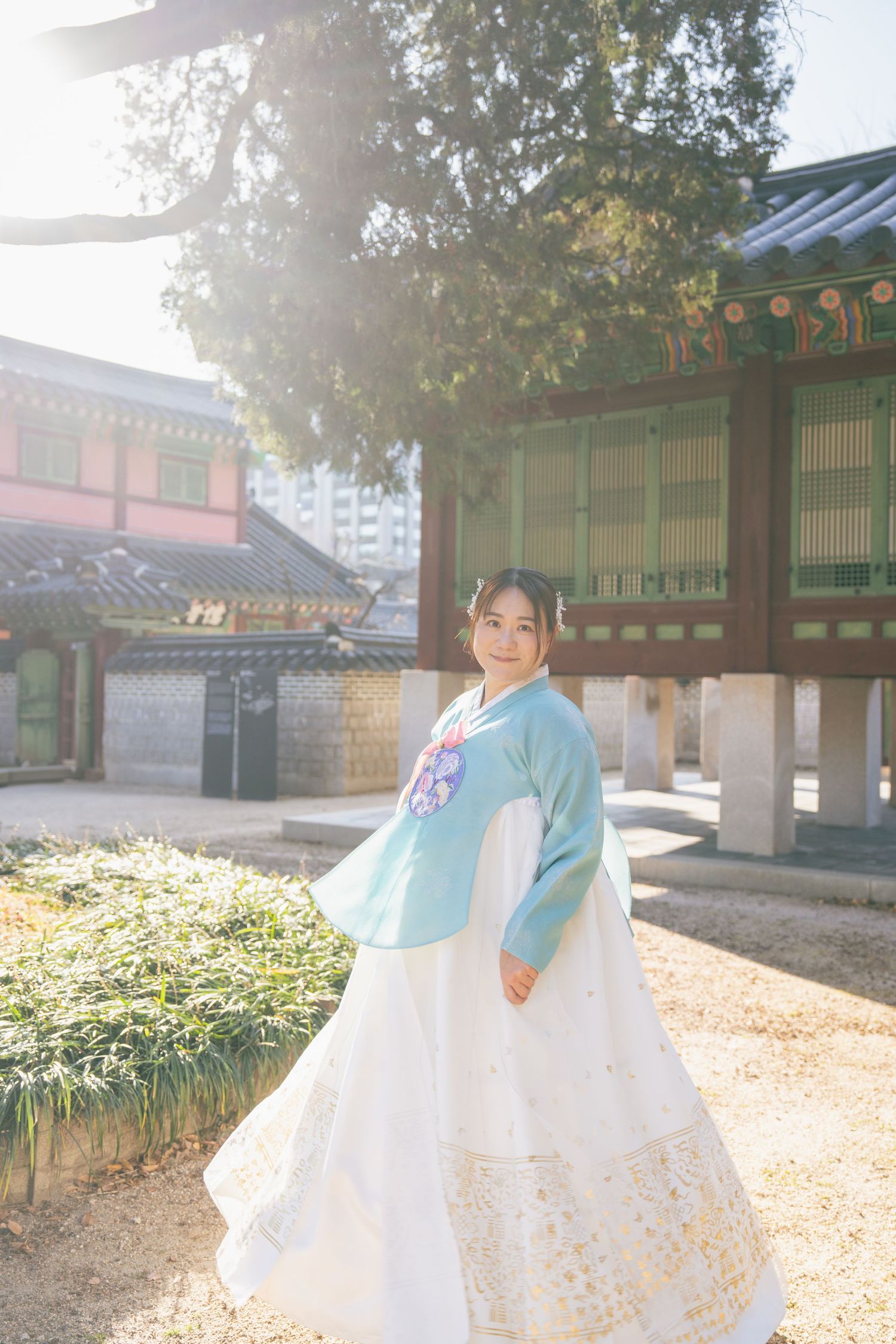 Person in traditional Korean hanbok dress poses gracefully in front of historic temple buildings on a sunny autumn day.