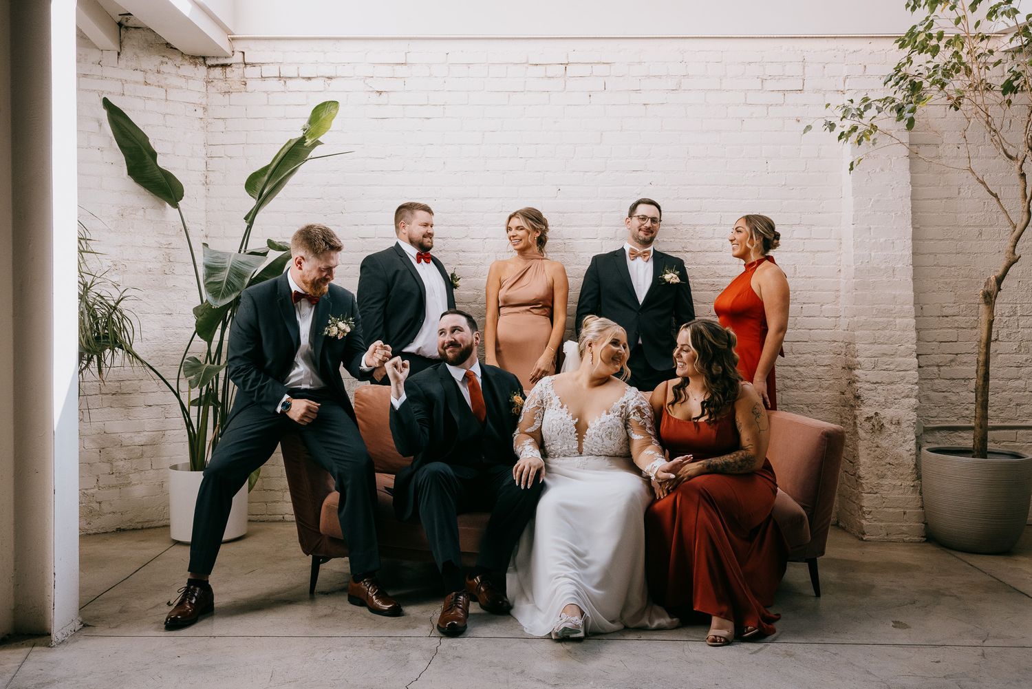 Candid modern wedding party at Gordon Green Lakewood Ohio, bride and groom on blush sofa with bridal party, white brick backdrop.