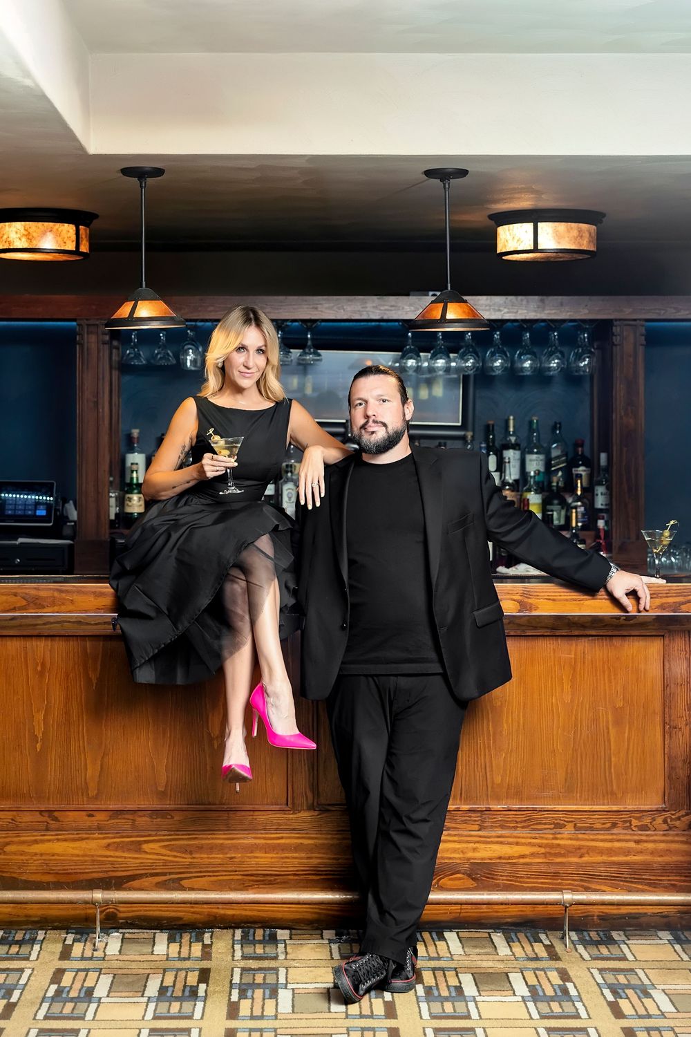 Stylish couple in black attire posing against wooden bar counter with pendant lighting overhead.