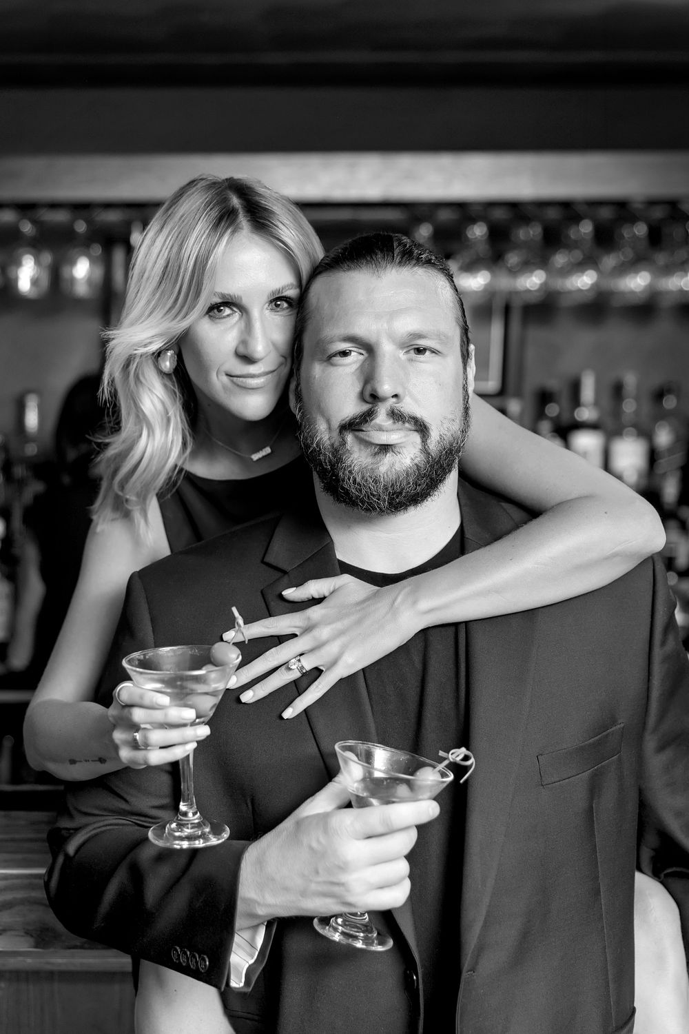 A couple poses together at a bar holding martini glasses in a black and white photograph.