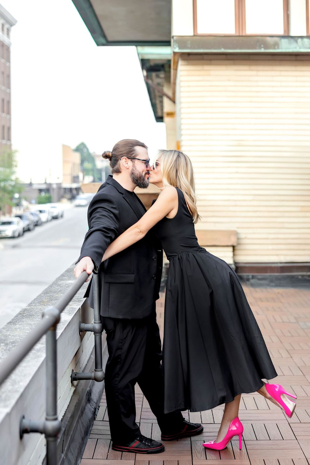 A romantic couple in matching black outfits embrace on a city balcony, with her bright pink heels adding a pop of color.