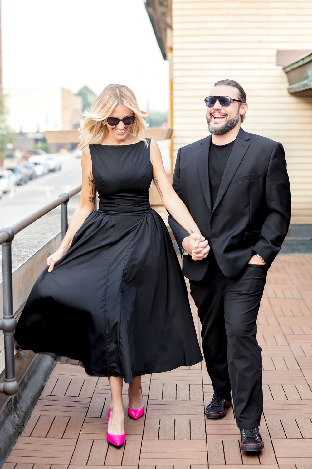 Couple in formal black outfits poses playfully on outdoor deck with pink heels.