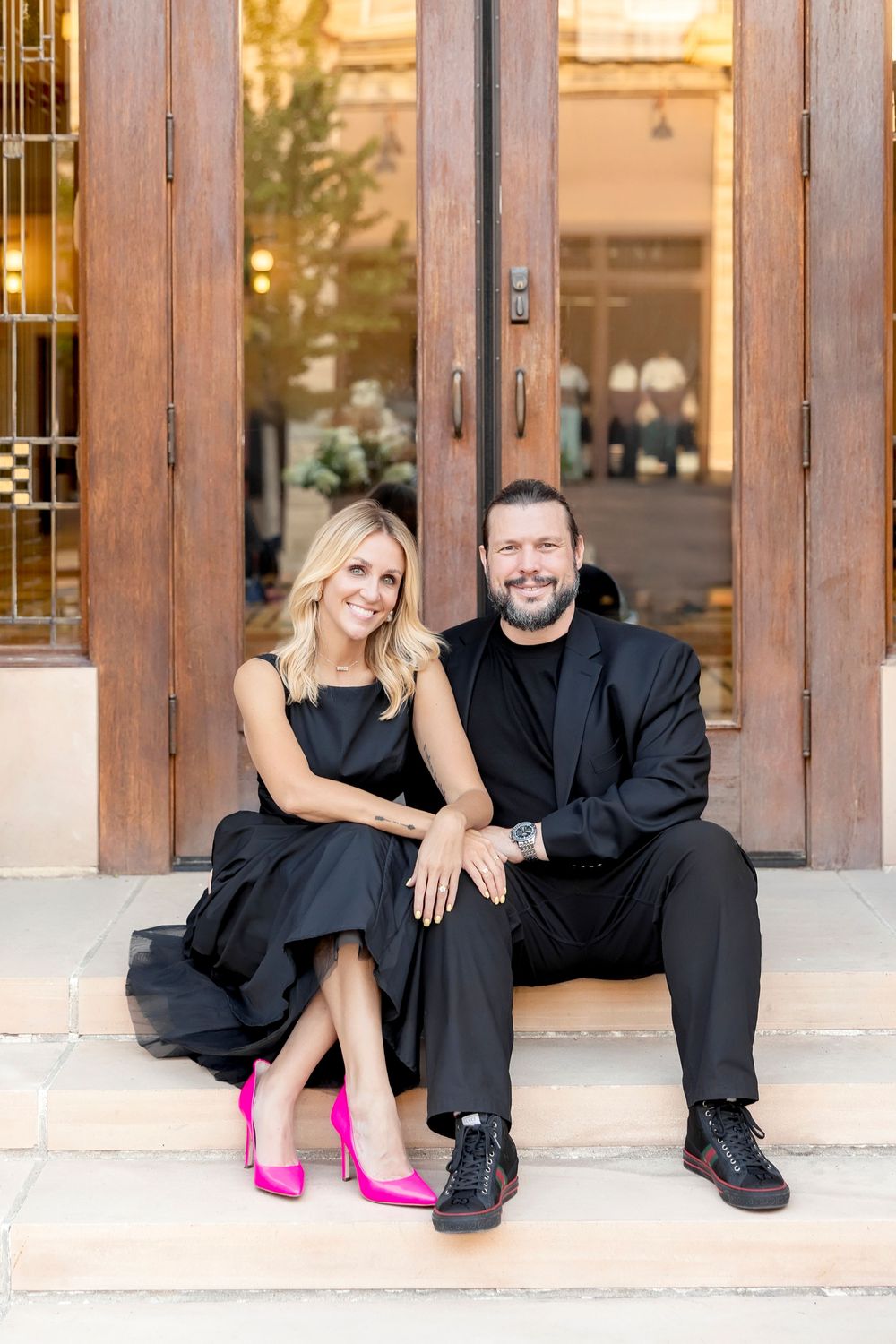Couple in black formal wear with pink heels sitting on wooden steps for romantic photoshoot.