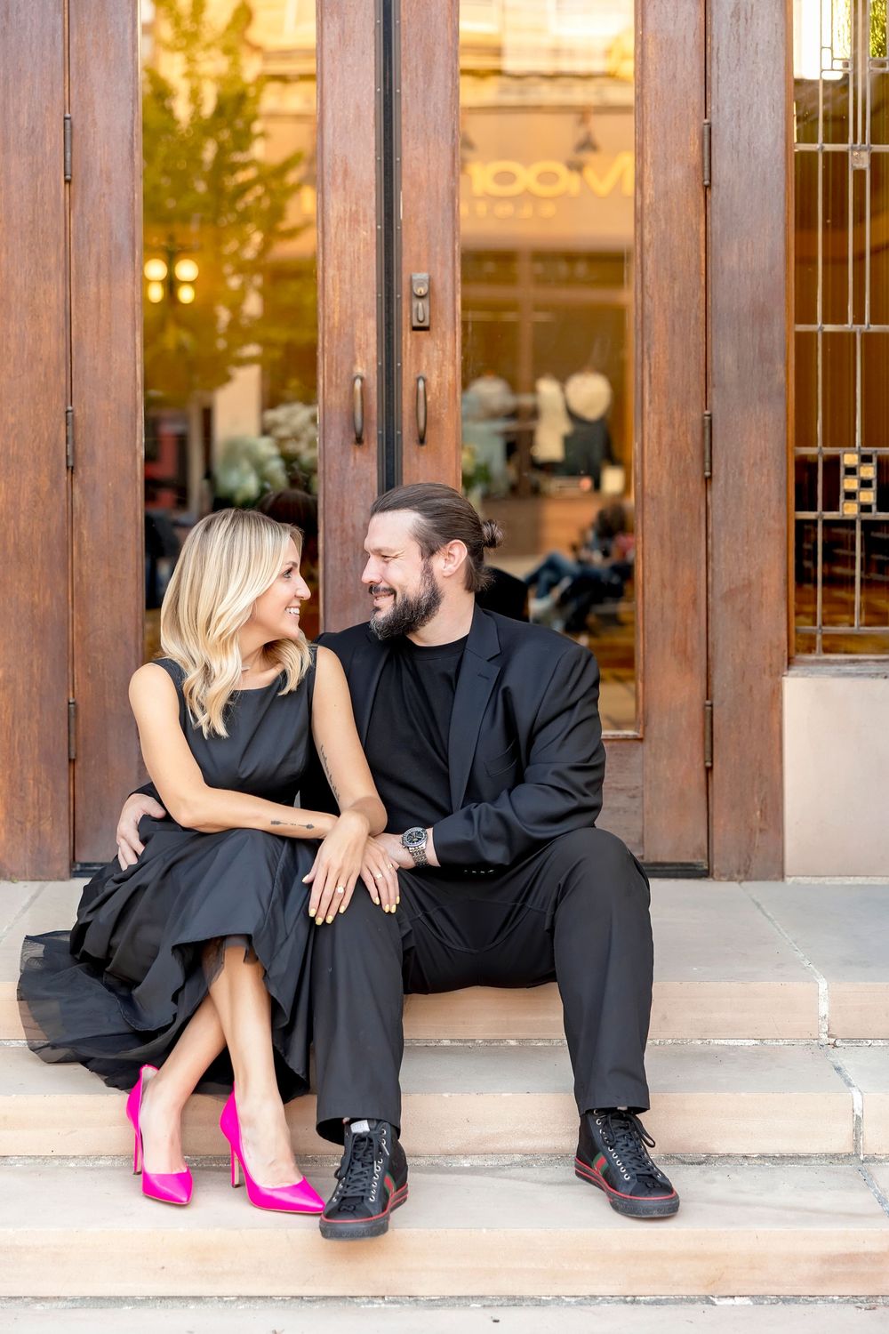 A couple in black attire sits on stone steps, with the woman wearing vibrant pink heels.