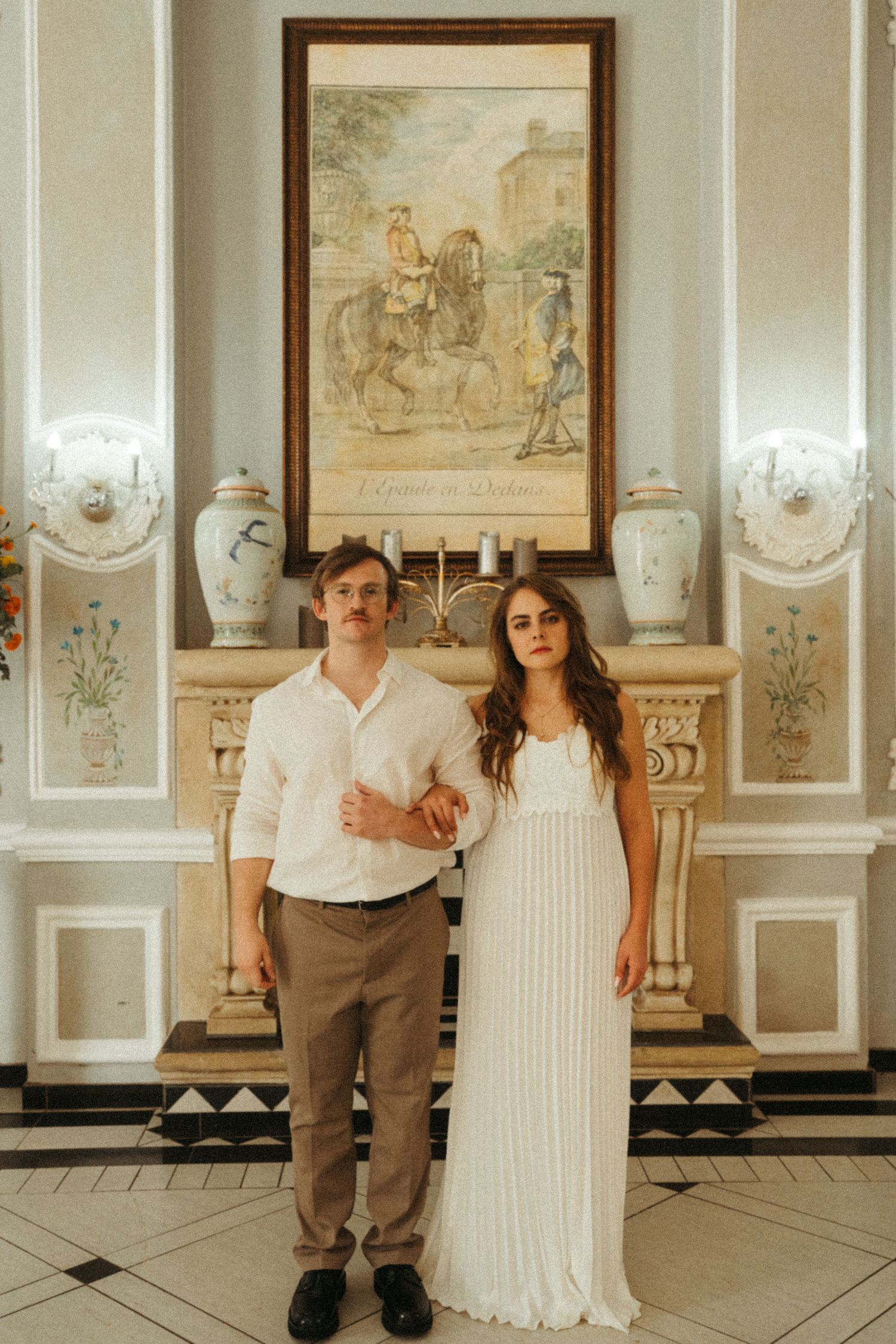 Two people stand together in a formal room with classical artwork and white wall panels.