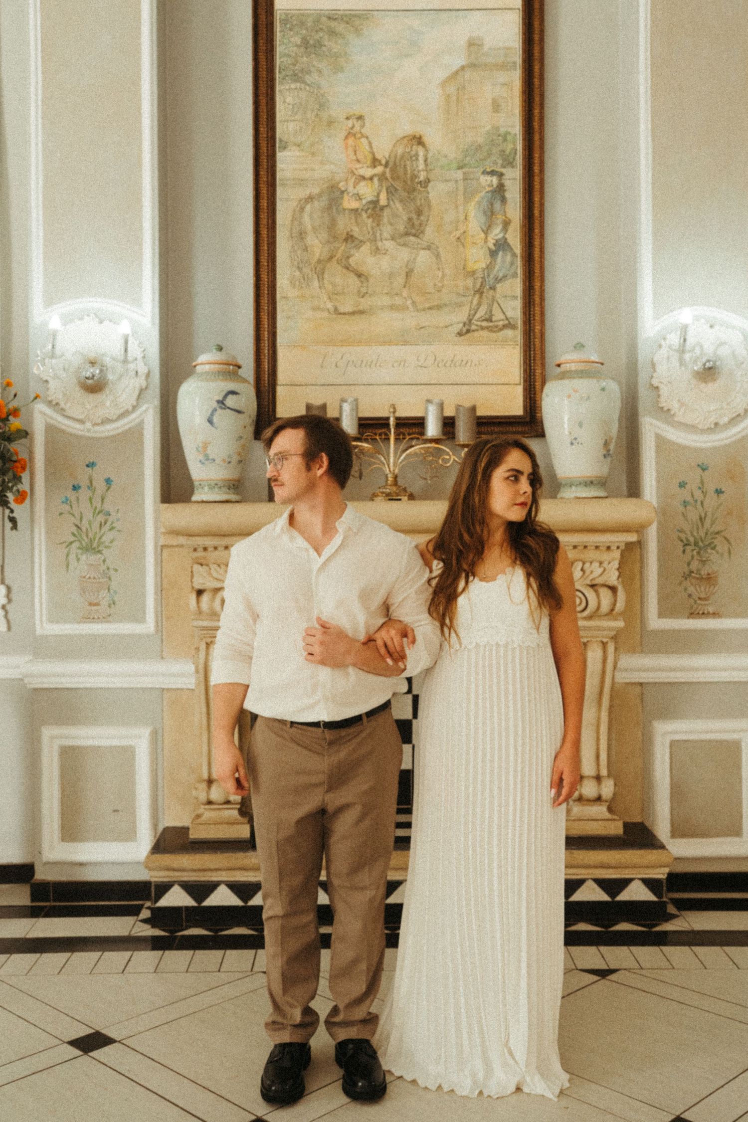 A couple stands side by side in an elegant white-paneled room with vintage artwork.