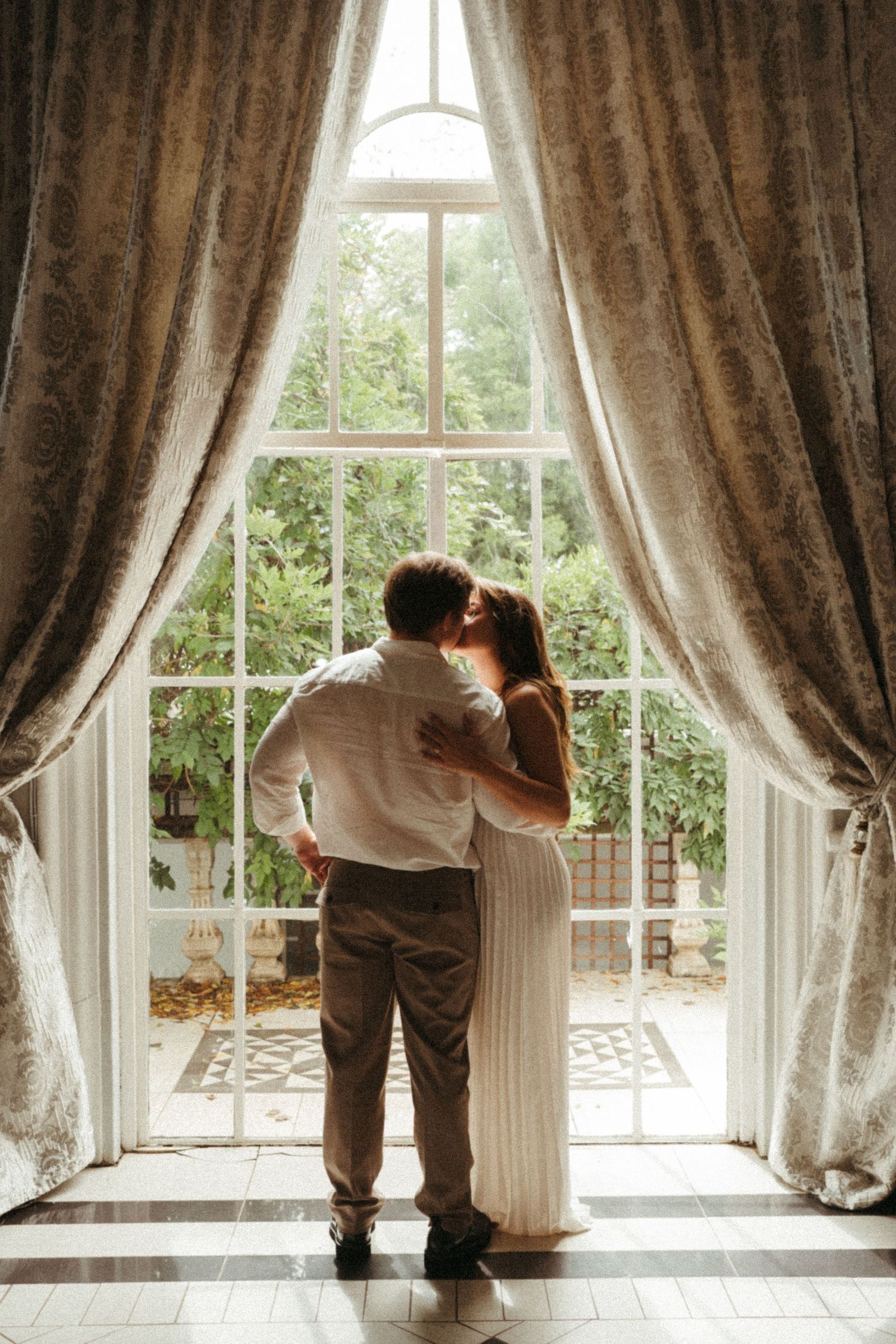 A couple shares an intimate moment by large windows with brown curtains.
