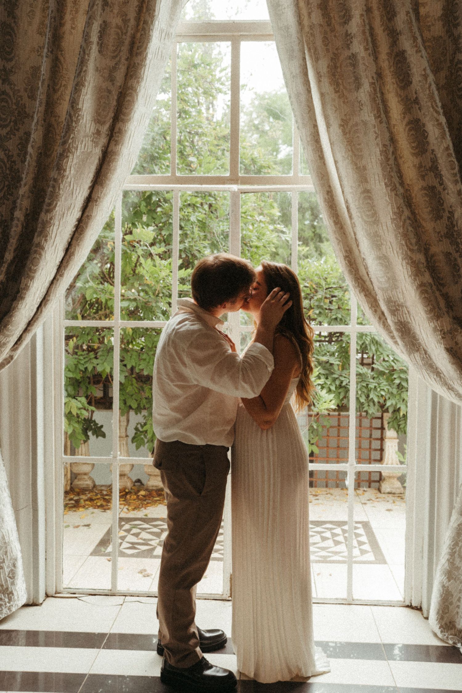 A romantic scene unfolds by large windows with flowing curtains and natural light.
