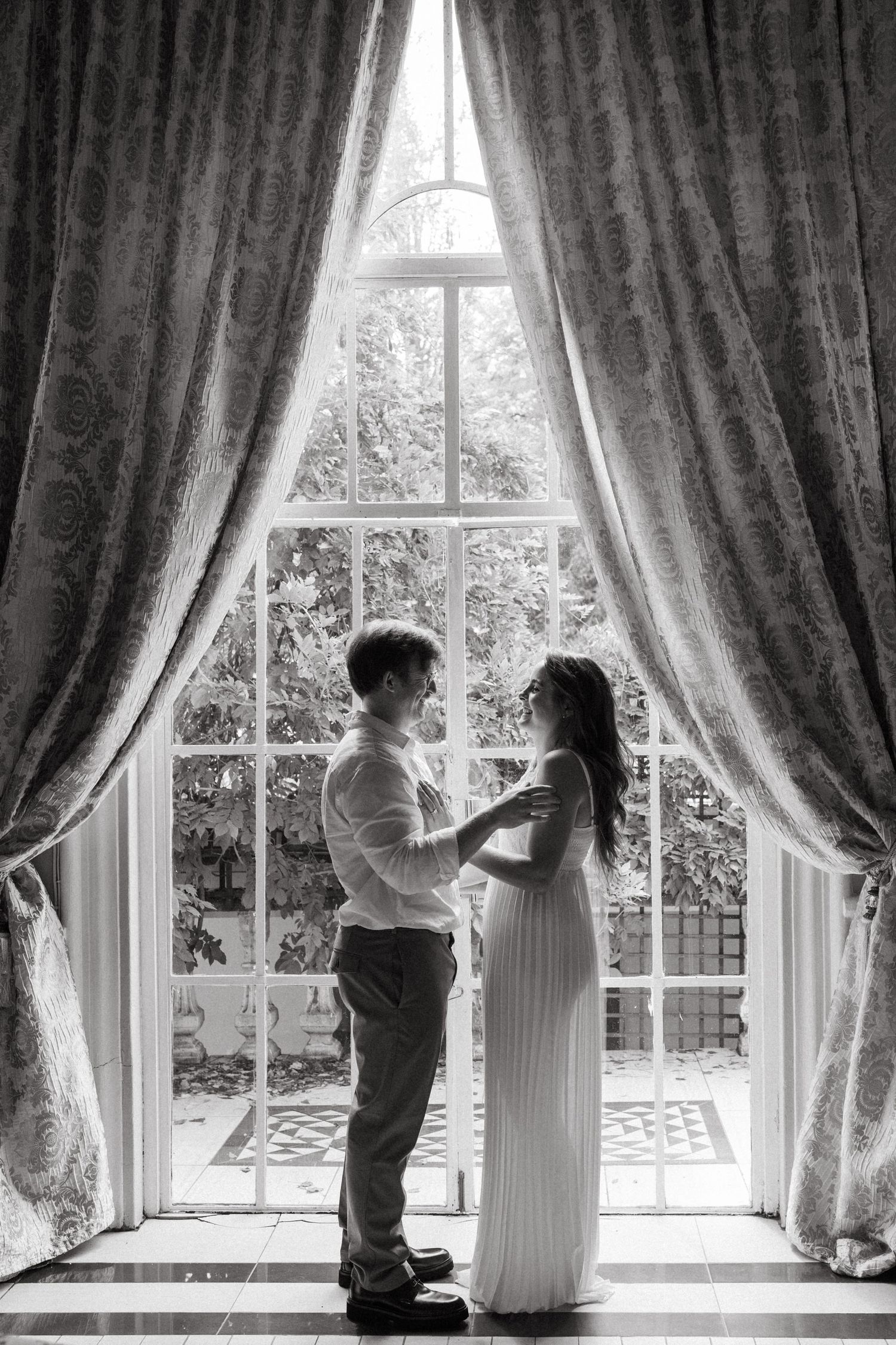 A tender moment captured in black and white by a window with elegant drapes.