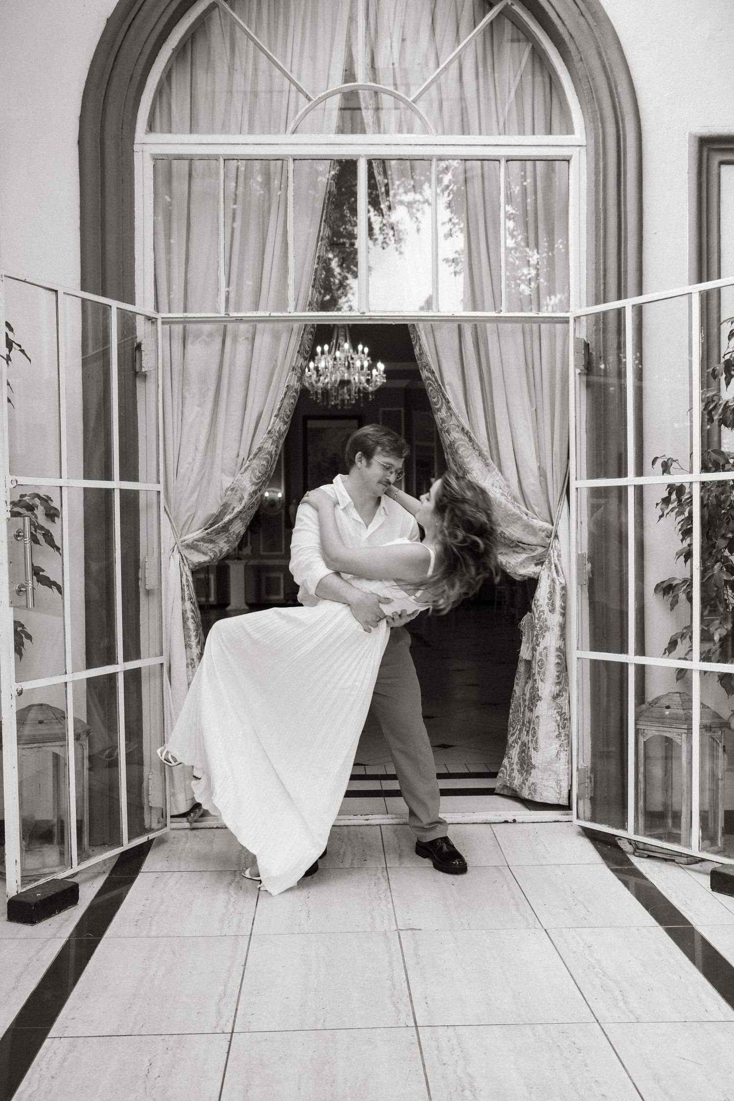 A romantic couple poses in an elegant indoor setting with arched windows and white curtains.