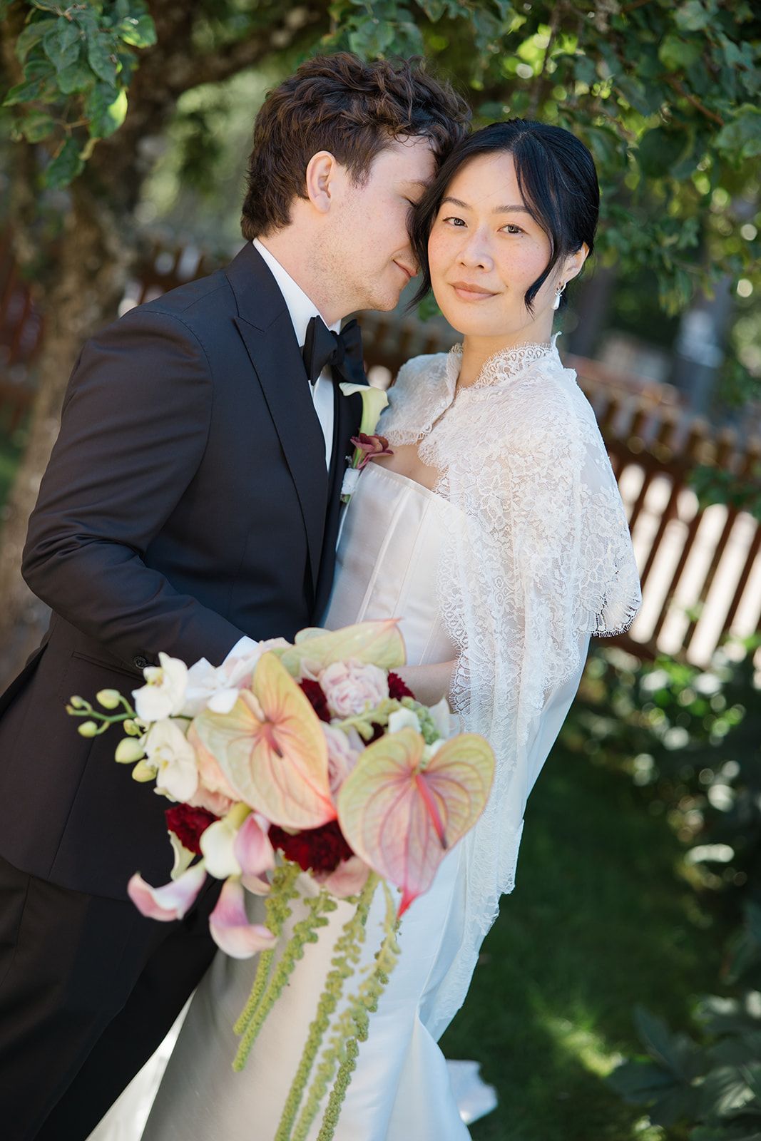 A romantic wedding couple shares tender moments in a garden setting while holding a pink and white floral bouquet.